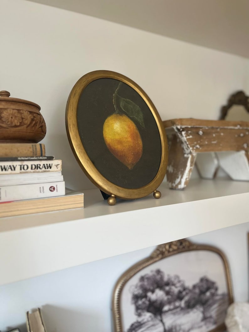 A lemon still life painting in a gold frame is on a shelf with antiques and vintage books.