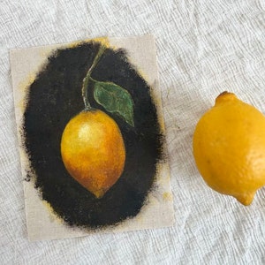 A still life vintage painting of a lemon is beside a real lemon on beige fabric.