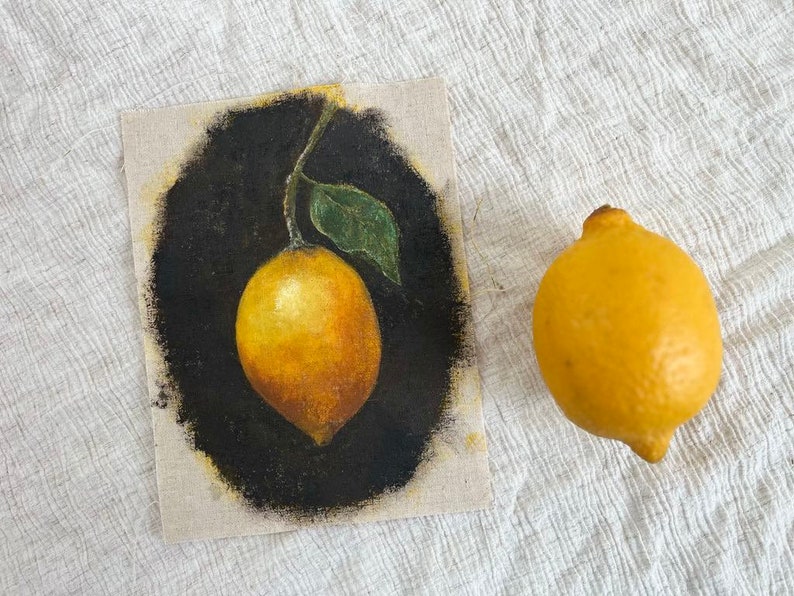 A still life vintage painting of a lemon is beside a real lemon on beige fabric.
