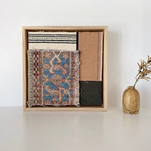 May include: A wooden frame holds a collection of fabric swatches, including a patterned rug, a striped fabric, and a solid brown fabric. The frame is on a white surface next to a small wooden vase with dried branches.