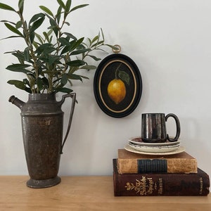 A lemon still life painting in a gold frame is hanging on a white wall above antiques and vintage books.