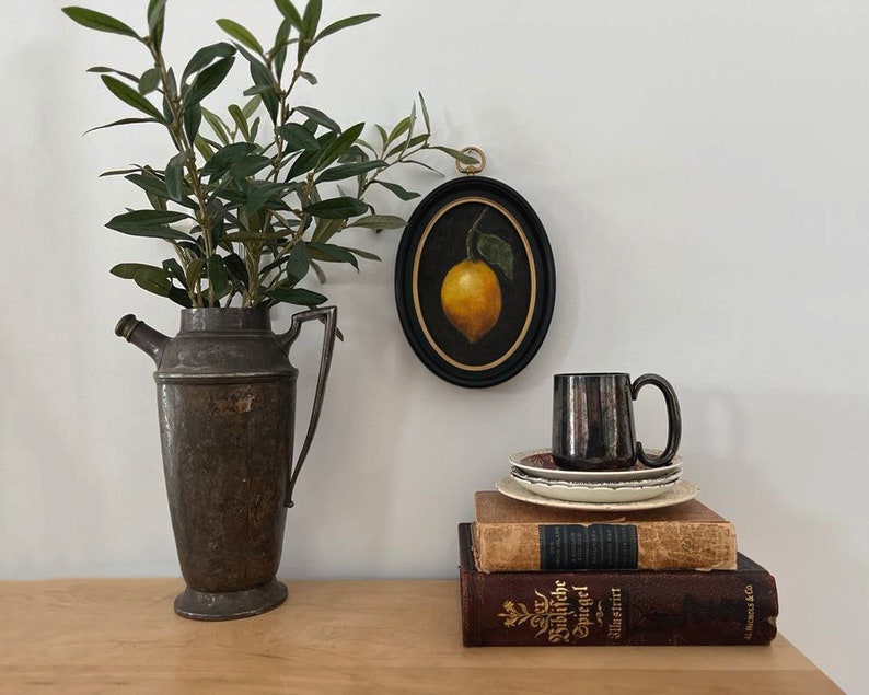 A lemon still life painting in a gold frame is hanging on a white wall above antiques and vintage books.