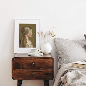 May include: A white framed print of a painting depicting a woman with long brown hair, wearing a white dress, against a green background. The print is on a wooden nightstand with a white ceramic vase and a white teacup and saucer.