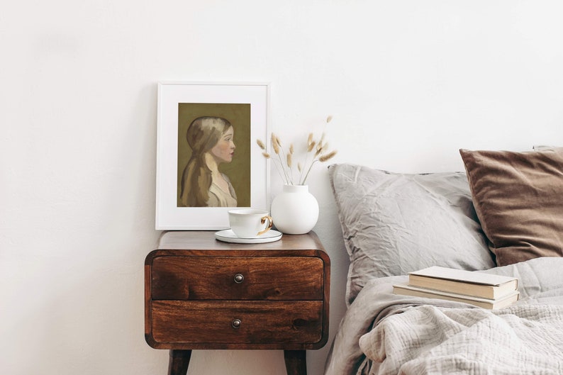 May include: A white framed print of a painting depicting a woman with long brown hair, wearing a white dress, against a green background. The print is on a wooden nightstand with a white ceramic vase and a white teacup and saucer.