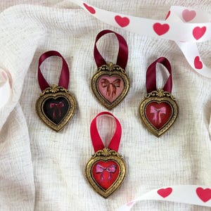 May include: Four gold heart-shaped frames with red and pink bows inside. The frames are hanging from red and burgundy ribbons on a white fabric background.
