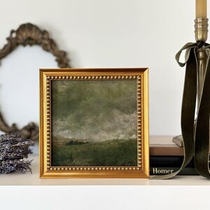 A gold frame with beading frames a vintage English countryside landscape art print. The frame is on a shelf with lavender, an antique mirror, vintage art books and a gold candle stick with a green velvet coquette bow.