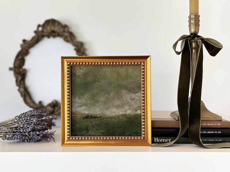 A gold frame with beading frames a vintage English countryside landscape art print. The frame is on a shelf with lavender, an antique mirror, vintage art books and a gold candle stick with a green velvet coquette bow.