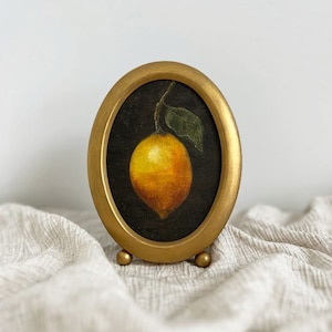 A lemon still life painting in a vintage oval gold frame is on beige fabric with a white wall behind it.