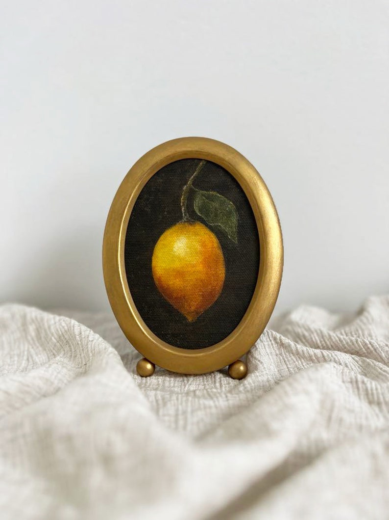 A lemon still life painting in a vintage oval gold frame is on beige fabric with a white wall behind it.