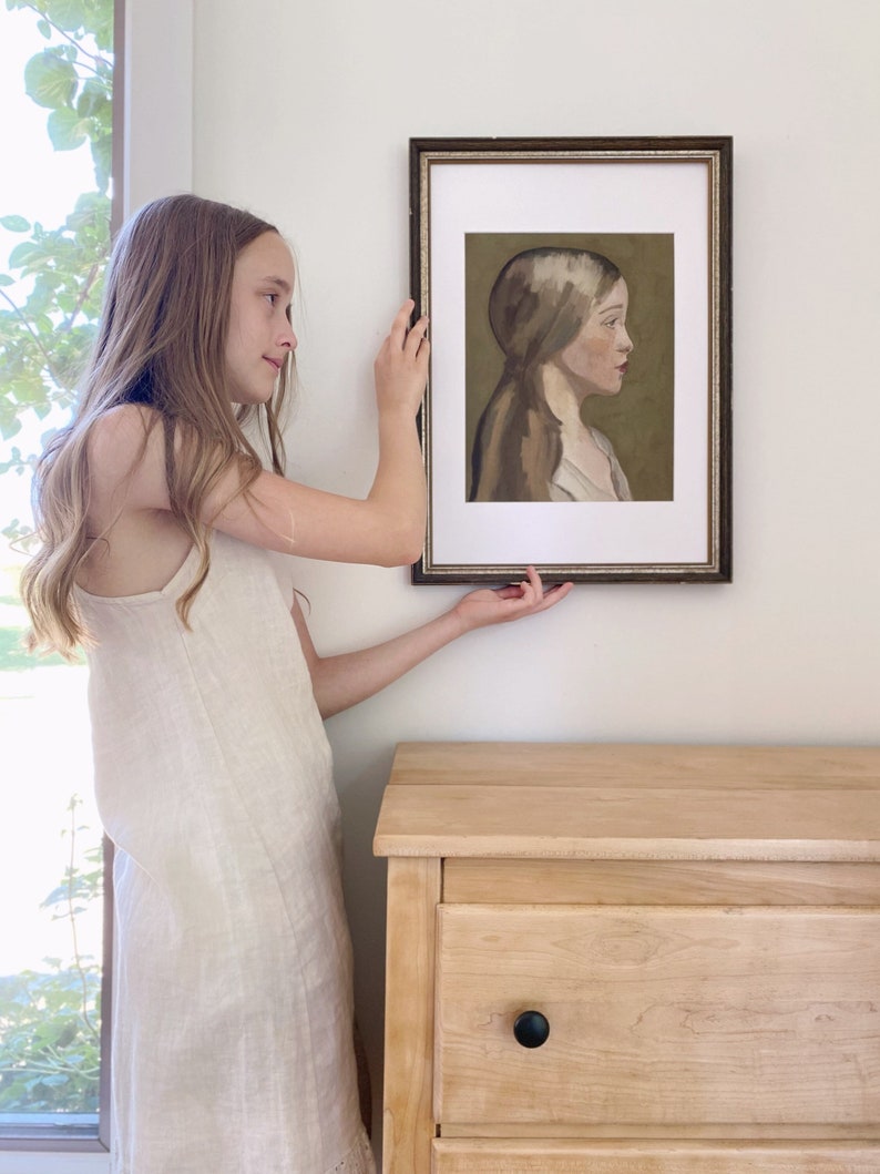 May include: A framed portrait of a young woman with long brown hair, wearing a white dress, against a green background. The portrait is hanging on a white wall.