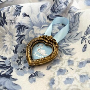 Tiny framed swan painting in a heart-shaped vintage gold frame with pale blue ribbon, displayed on blue and white roses, romantic cottagecore decor.