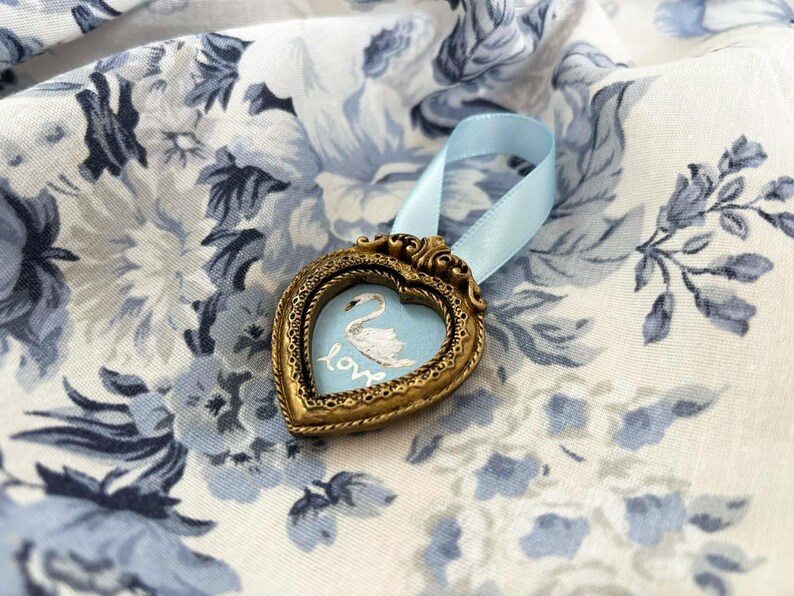 Tiny framed swan painting in a heart-shaped vintage gold frame with pale blue ribbon, displayed on blue and white roses, romantic cottagecore decor.