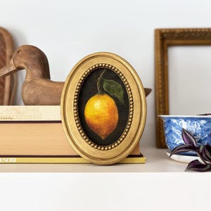 A lemon still life painting in a gold oval frame is on a shelf with antiques and vintage books and a plant in a blue chintz planter.