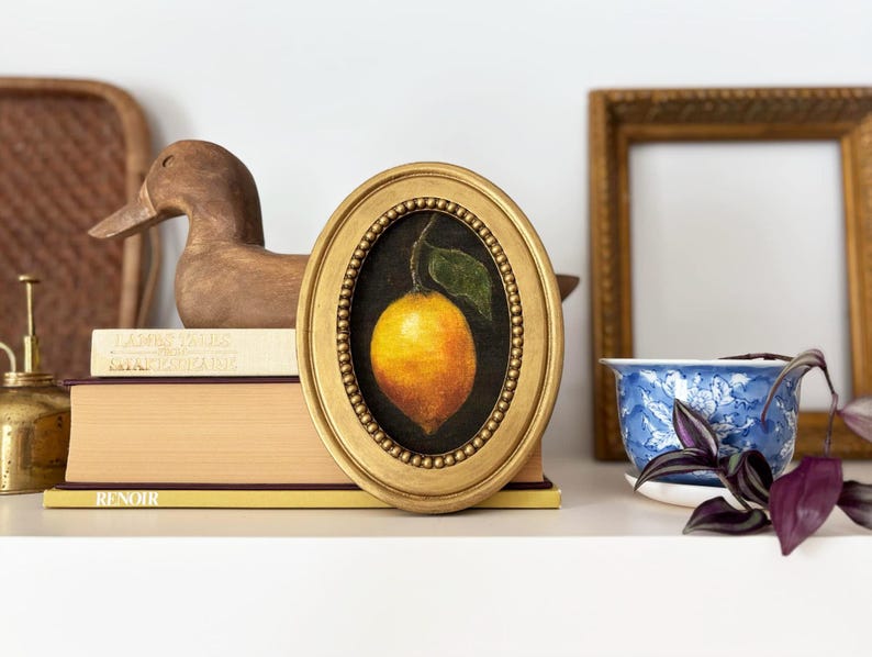 A lemon still life painting in a gold oval frame is on a shelf with antiques and vintage books and a plant in a blue chintz planter.