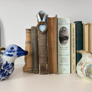 Tiny framed swan painting in a heart-shaped vintage gold frame with pale blue ribbon, displayed on a shelf with vintage books, a vintage-style blue and white English China duck and romantic cottagecore decor.