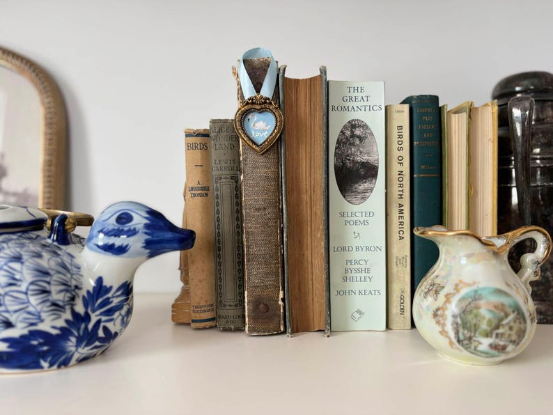 Tiny framed swan painting in a heart-shaped vintage gold frame with pale blue ribbon, displayed on a shelf with vintage books, a vintage-style blue and white English China duck and romantic cottagecore decor.