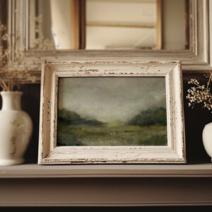 A shabby chic creamy wooden frame holds a vintage landscape oil painting on a mantle beside dried flowers in pots.