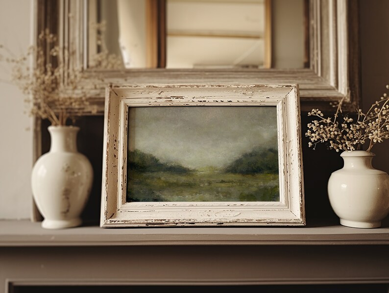 A shabby chic creamy wooden frame holds a vintage landscape oil painting on a mantle beside dried flowers in pots.