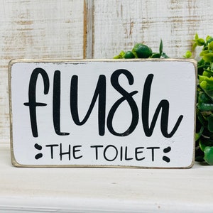 May include: White wooden sign with black text that reads "Flush the Toilet".