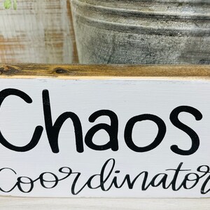 Puede incluir: Un letrero rectangular de madera con una parte frontal pintada de blanco y una parte superior de madera natural. El letrero dice "Chaos Coordinator" en letras negras y en escritura. El letrero es un art&iacute;culo decorativo.