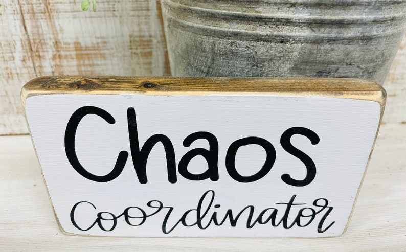 Puede incluir: Un letrero rectangular de madera con una parte frontal pintada de blanco y una parte superior de madera natural. El letrero dice "Chaos Coordinator" en letras negras y en escritura. El letrero es un art&iacute;culo decorativo.