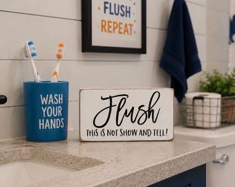 May include: A rectangular white wooden sign with black text that reads Flush THIS IS NOT SHOW AND TELL, positioned next to a blue cylindrical toothbrush holder with the white text WASH YOUR HANDS and two toothbrushes inside the holder.