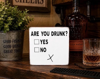 Może przedstawiać: Kwadratowy biały drewniany znak z przetartymi krawędziami, zawierający czarny napis ARE YOU DRUNK? oraz dwa pola wyboru oznaczone jako YES i NO, z odręcznie narysowanym czarnym znakiem X umieszczonym poniżej pól na białej powierzchni.