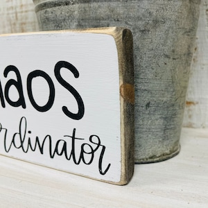 Puede incluir: Un letrero rectangular de madera con las palabras "Chaos Coordinator" en escritura negra sobre un fondo blanco. El letrero tiene un aspecto r&uacute;stico y desgastado. Un cubo de metal gris en el fondo.