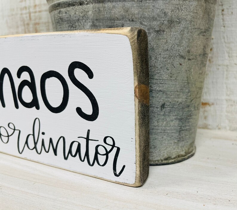 Puede incluir: Un letrero rectangular de madera con las palabras "Chaos Coordinator" en escritura negra sobre un fondo blanco. El letrero tiene un aspecto r&uacute;stico y desgastado. Un cubo de metal gris en el fondo.