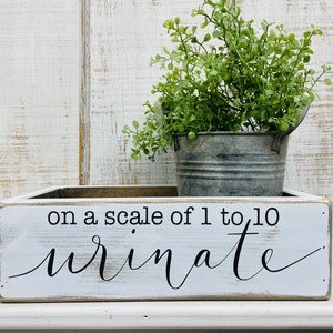 May include: A rectangular white wooden box with distressed edges featuring the black text on a scale of 1 to 10 urinate. A small galvanized metal bucket containing a green artificial plant sits inside the wooden box on a white surface.