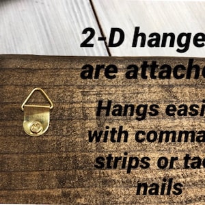 May include: A close-up of a wooden sign with the text "2-D hangers are attached. Hangs easily with command strips or tack nails." The sign has two gold-colored D-shaped hangers on the back.