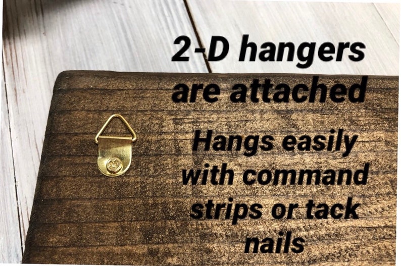 May include: A close-up of a wooden sign with the text "2-D hangers are attached. Hangs easily with command strips or tack nails." The sign has two gold-colored D-shaped hangers on the back.