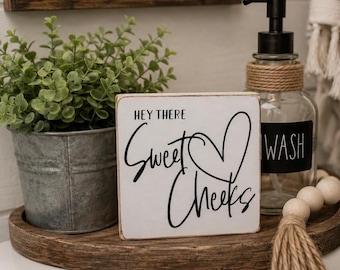 May include: A square white wooden sign with black text that reads "HEY THERE Sweet Cheeks" and a black heart outline. The sign is positioned on a dark brown wooden tray next to a small galvanized metal bucket with a green plant and a glass soap dispenser.