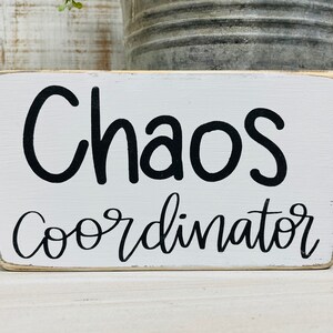 Puede incluir: Un letrero rectangular de madera blanca con las palabras "Chaos Coordinator" en escritura negra. El letrero tiene un aspecto envejecido y vintage, con bordes ligeramente desgastados. Un cubo de metal y vegetaci&oacute;n son visibles en el fondo.
