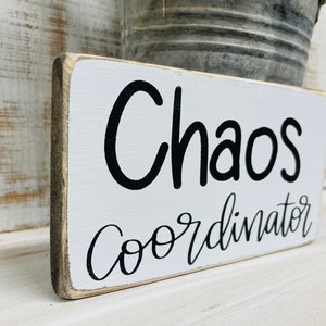 Puede incluir: Un letrero rectangular de madera con las palabras "Chaos Coordinator" en letras negras sobre un fondo blanco. El letrero tiene un aspecto envejecido y r&uacute;stico con un borde gris.
