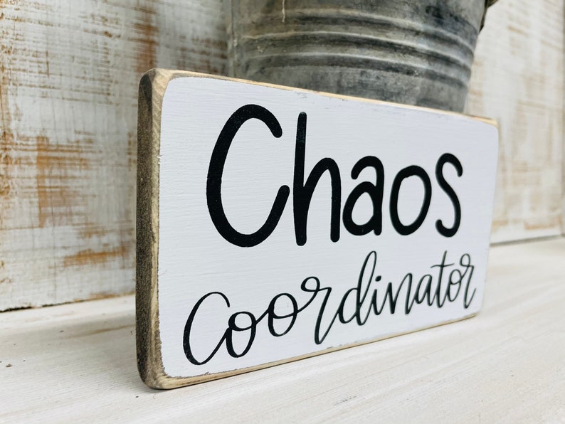 Puede incluir: Un letrero rectangular de madera con las palabras "Chaos Coordinator" en letras negras sobre un fondo blanco. El letrero tiene un aspecto envejecido y r&uacute;stico con un borde gris.