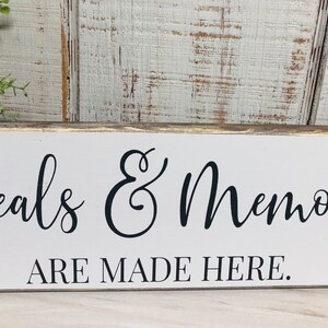 May include: White wooden sign with black text that reads "Meals & Memories Are Made Here."