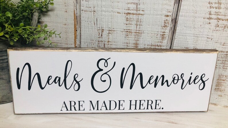 May include: White wooden sign with black text that reads "Meals & Memories Are Made Here."