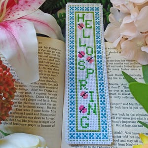 May include: A white bookmark with a blue and green border. The bookmark is embroidered with the words "Hello Spring" in green and pink thread. There are small ladybugs embroidered in pink and green thread.
