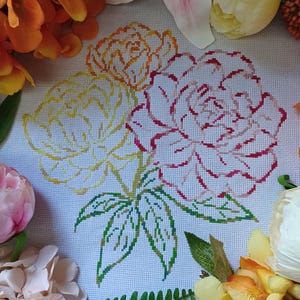 May include: A cross-stitch floral design featuring three roses in shades of yellow, orange, and red, with green leaves. The embroidery is set against a white background, surrounded by artificial flowers in various colors.