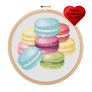 May include: A cross stitch pattern featuring a colorful assortment of macarons in a hoop. The macarons are in shades of blue, green, yellow, pink, and red.