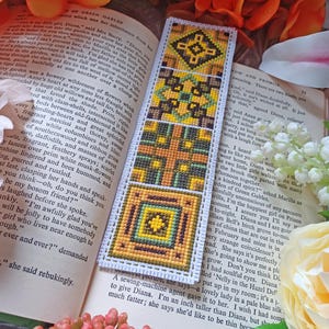 May include: A bookmark with a geometric pattern in shades of yellow, green, and brown. The bookmark is made of fabric and has a fringed edge.