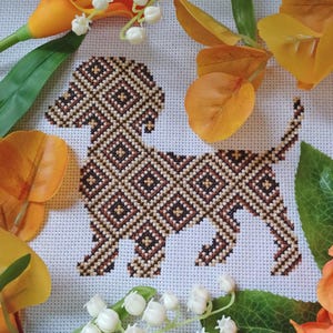 Puede incluir: Patrón de punto de cruz de un perro salchicha marrón y beige sobre tela blanca rodeado de flores naranjas y blancas.