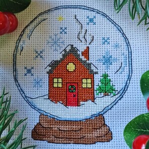 May include: Cross-stitch pattern of a snow globe with a red house, a green tree, and snowflakes inside. The snow globe is on a brown base.