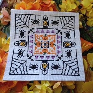 May include: A white cross-stitch pattern with a Halloween theme. The center features a repeating pattern of orange pumpkins and purple witch hats. The corners feature black spiders and orange jack-o'-lanterns.