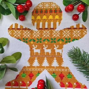 Puede incluir: Un patrón de punto de cruz de un hombre de jengibre que lleva un suéter festivo con renos, árboles de Navidad y adornos. El hombre de jengibre es marrón con detalles de glaseado blanco y un gorro rojo y verde.