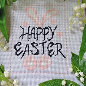 May include: A white cross-stitch embroidery with pink bunny ears and pink eggs. The text "Happy Easter" is stitched in black thread.