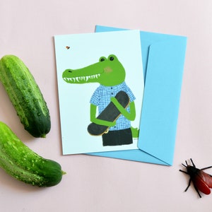 Carte Postale Crocodile skateur. Illustration de milicancrelat d'un croco vert tenant sa planche de skate. Verso imprimé. 10,5x14,8 cm