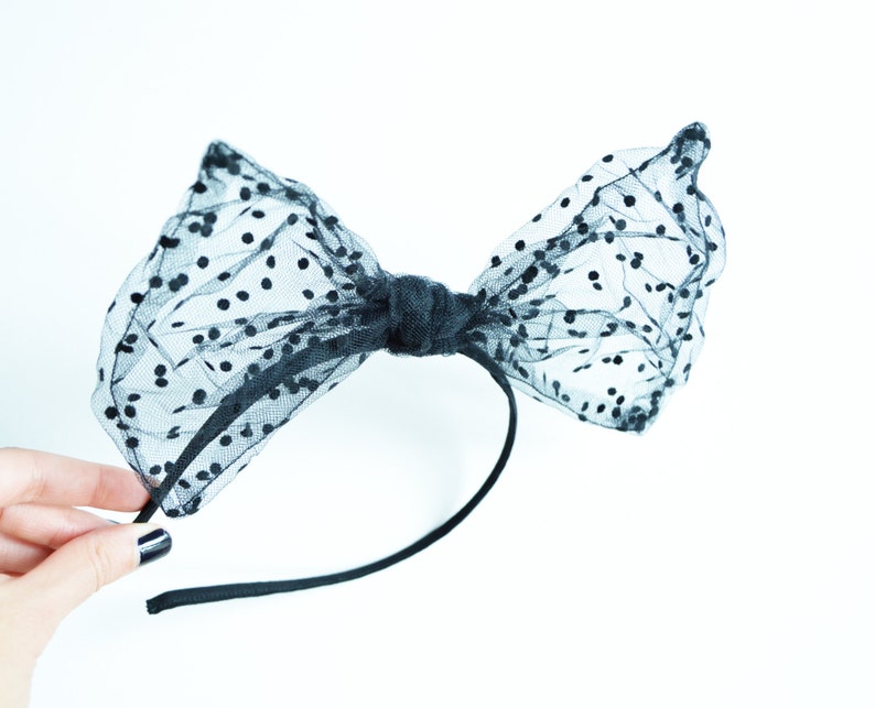 May include: Black polka dot bow headband. The bow is made of sheer black fabric with small, evenly spaced black dots. The bow is tied in the center and attached to a thin black headband. The accessory is designed for hair styling.