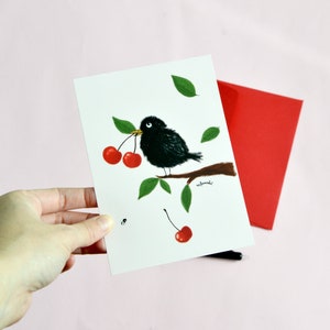 Carte Postale petit oiseau noir sur sa branche picorant des cerises sur fond rose pâle.  Carte 14,8 cm X 10,5 cm.